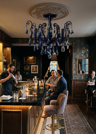 A dimly lit upscale bar interior with dark wood cabinetry, a large blue ornate chandelier, a bar counter with stools, and several people seated or standing in the space.