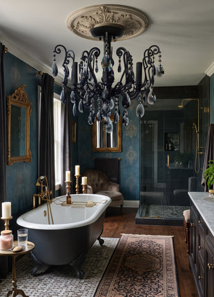 A luxurious bathroom interior with deep teal patterned wallpaper, a black clawfoot bathtub, a large ornate black chandelier, gold-framed mirrors, and a double vanity with a white marble countertop.