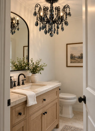 An elegant bathroom vanity area featuring a black metal chandelier with hanging crystal drops, a light wood double-sink vanity with a marble countertop, a white toilet, and framed wall art.