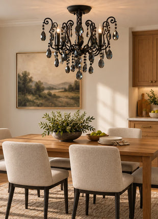 A styled dining room scene featuring a rustic wooden dining table with six upholstered dining chairs, a black metal chandelier with hanging glass teardrop crystals, a landscape painting on the wall, and a wooden kitchen cabinet unit in the background.