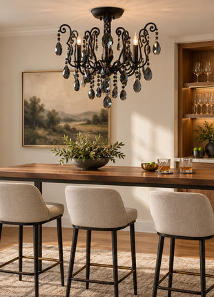 A styled interior dining space with a dark wood bar-height table, four upholstered bar stools, a black ornate chandelier, a framed landscape painting, and a wood shelving unit in the background.
