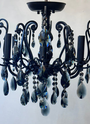 A black wrought-iron style chandelier with multiple teardrop-shaped dark blue glass crystal drops and candle-shaped bulb holders, shown against a plain light gray wall.
